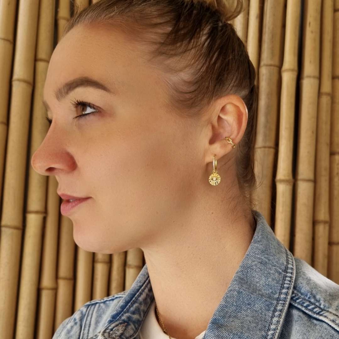 Model draagt vergulde creolen met hanger in de vorm van een maya oogje met gekleurde steentjes